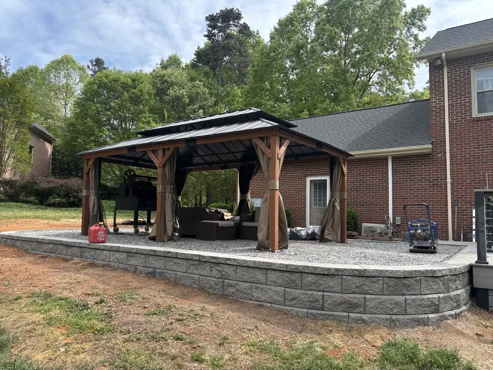 Raised gravel pad held by a block retaining wall, with a gazebo set on top, behind a brick house