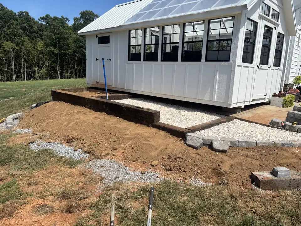 Landscape timbers framing white gravel beside a greenhouse, with block edging and graded dirt